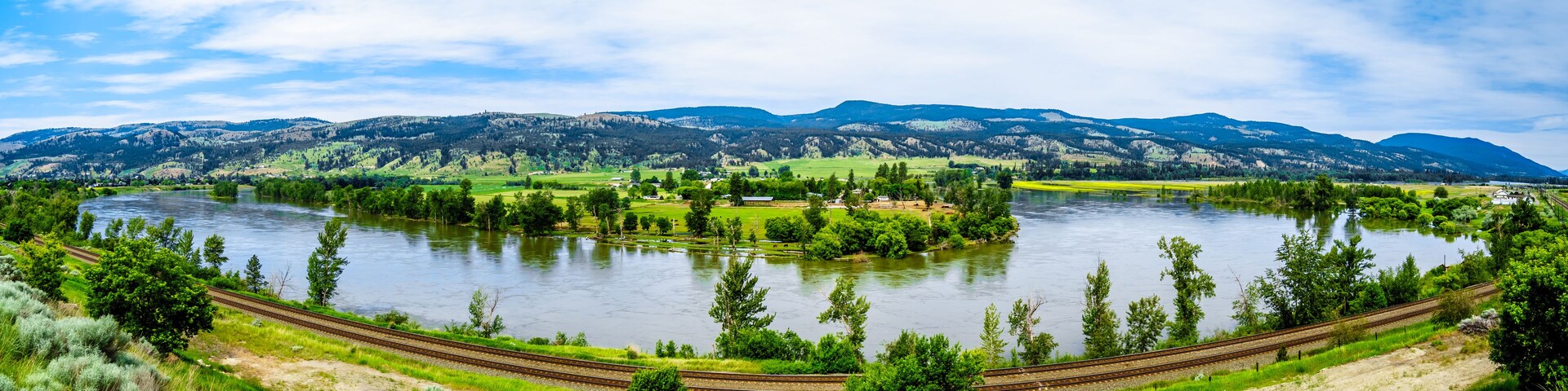Un río con una vía de tren que serpentea a su alrededor, rodeado de campos verdes y montañas al fondo.