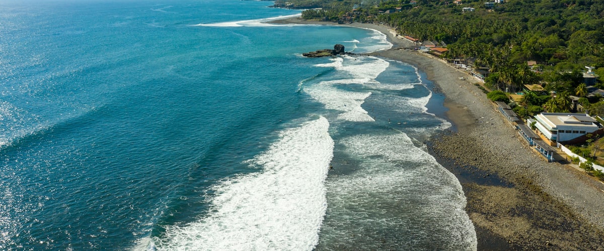 A coastal landscape with a clear blue sea, a sandy beach, and a hilly terrain with buildings.