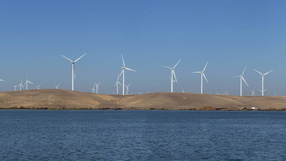 A landscape with multiple wind turbines on a hill by a body of water.