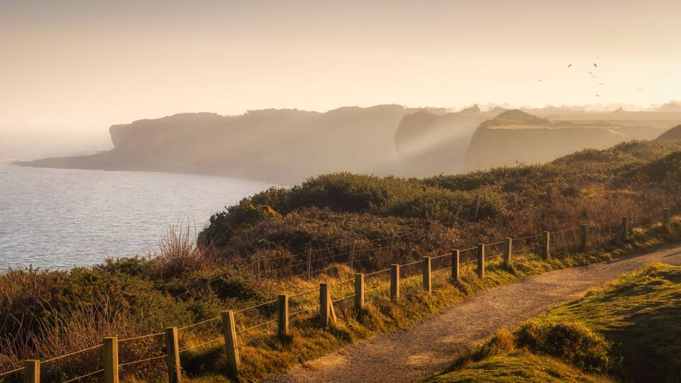 A coastal landscape with a dirt path, a wooden fence, and cliffs overlooking the sea.