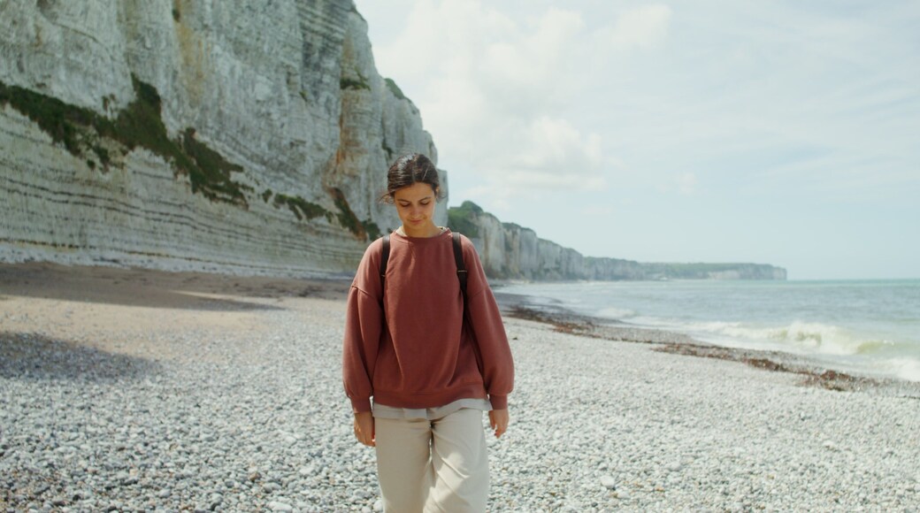 A young beautiful woman walks along a pebbly beach past sheer limestone cliffs, admiring the picturesque seascape. Etretat, France. 4k video, red komodo