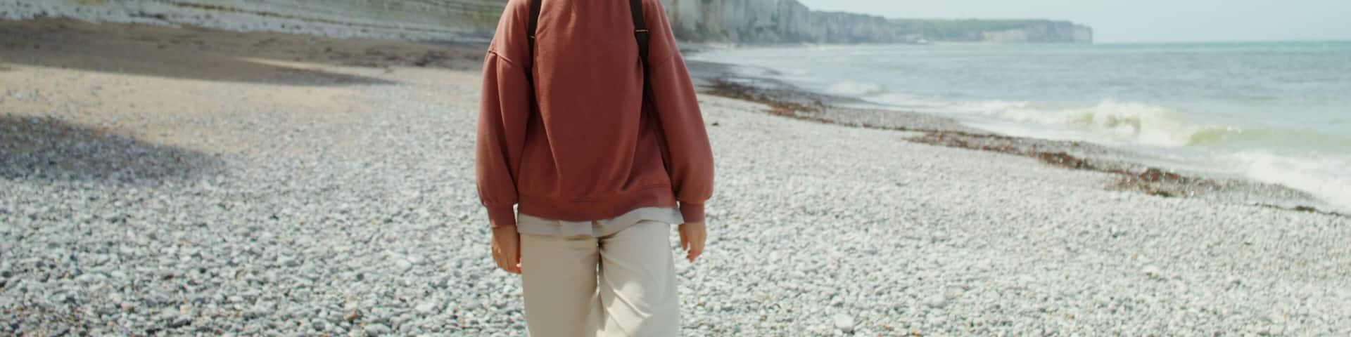 A young beautiful woman walks along a pebbly beach past sheer limestone cliffs, admiring the picturesque seascape. Etretat, France. 4k video, red komodo