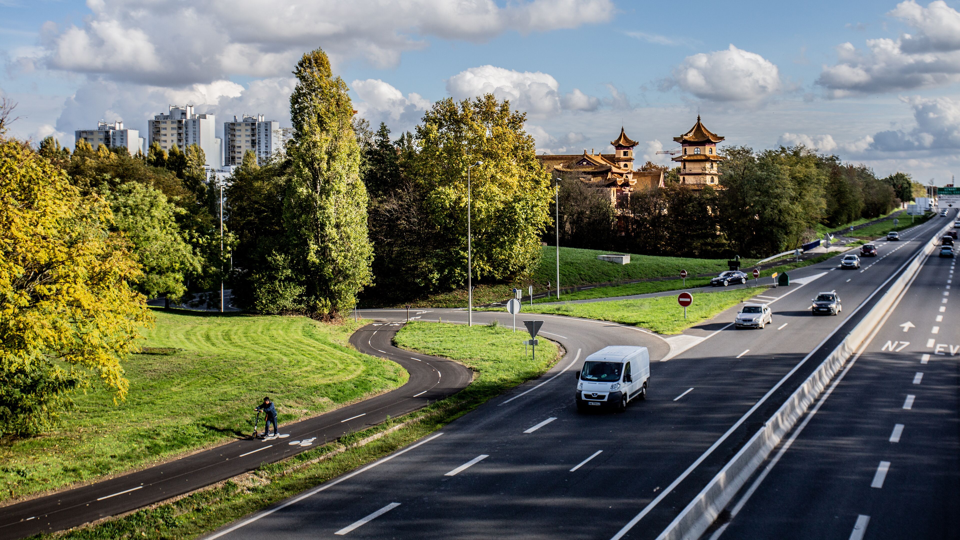 A highway with a curve, a van, and a scooter. There are trees, a park, and a traditional building in the background.