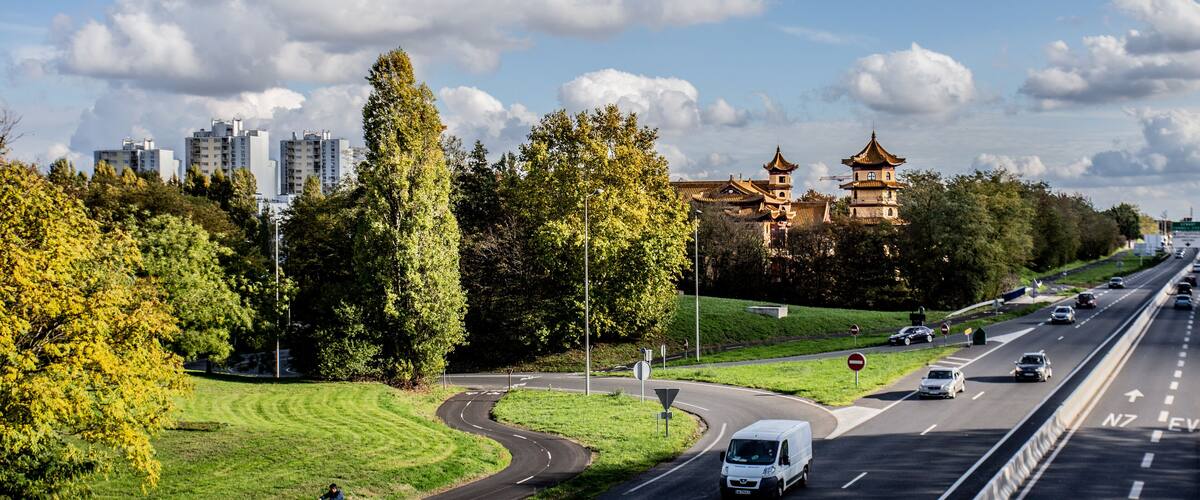 A highway with a curve, a van, and a scooter. There are trees, a park, and a traditional building in the background.