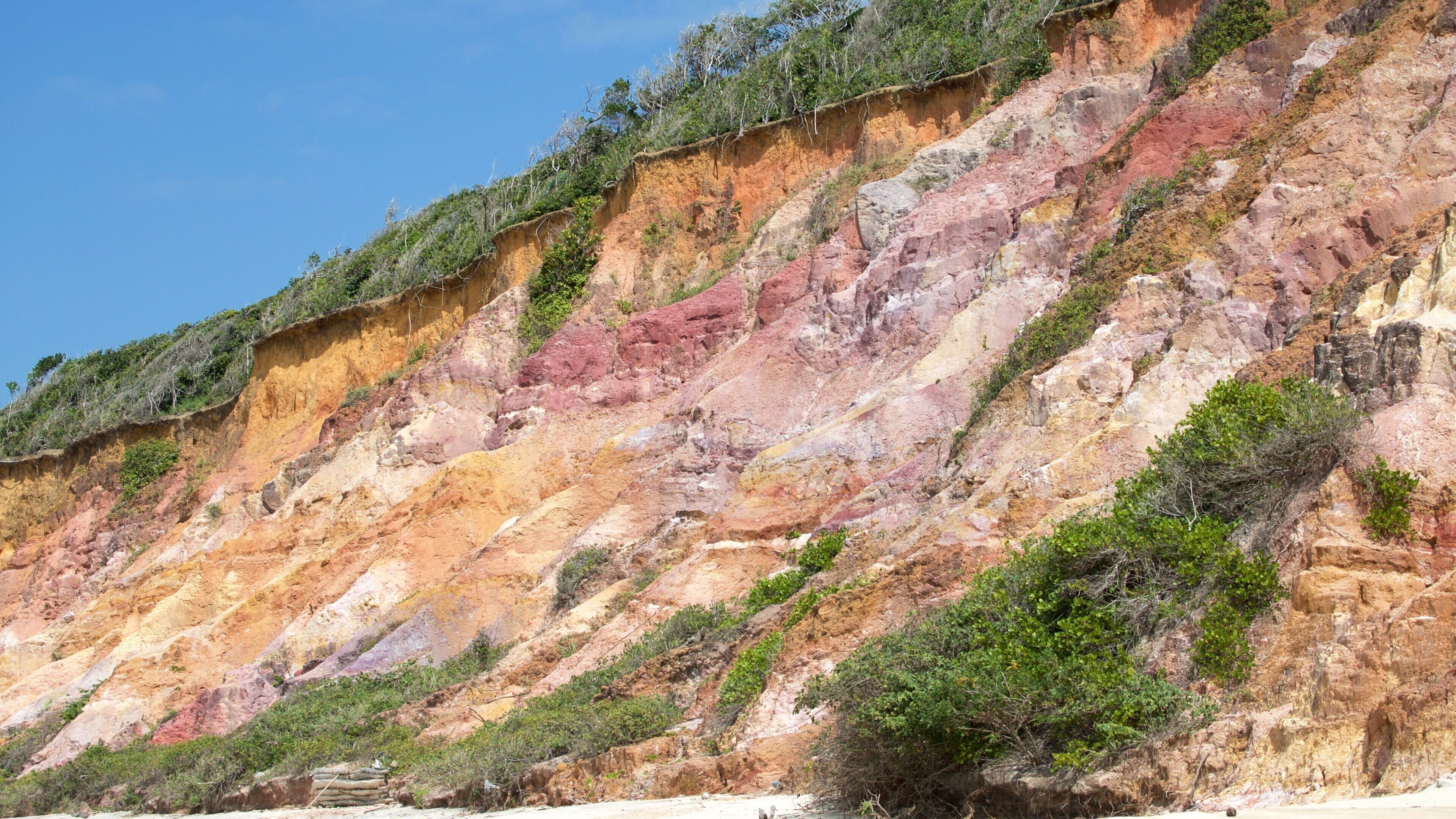 A colorful cliff with vegetation at the top and a sandy beach at the bottom.