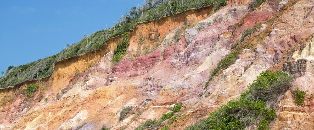 A colorful cliff with vegetation at the top and a sandy beach at the bottom.