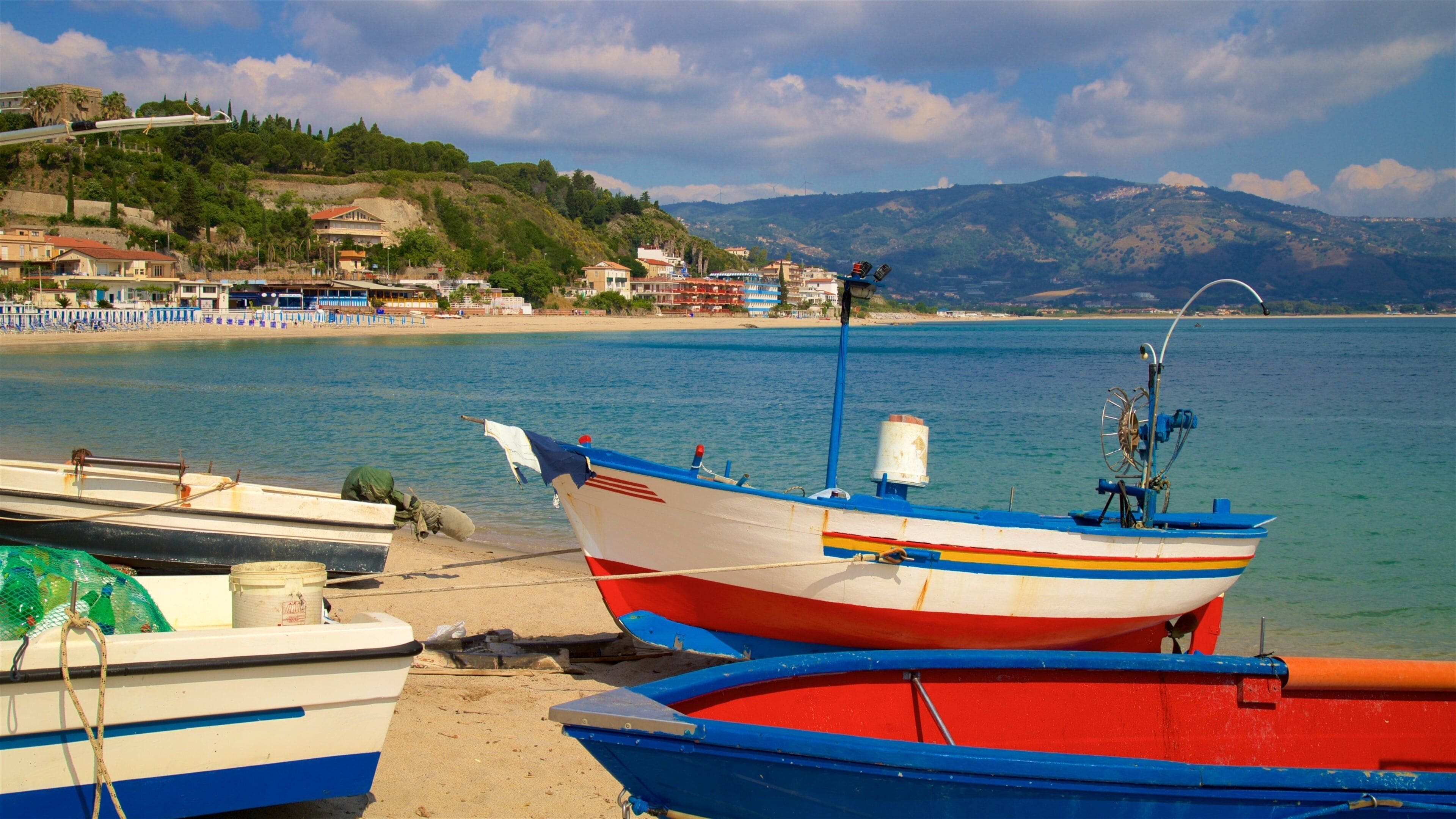Un paisaje costero con barcos amarrados en la orilla, una playa y montañas al fondo.