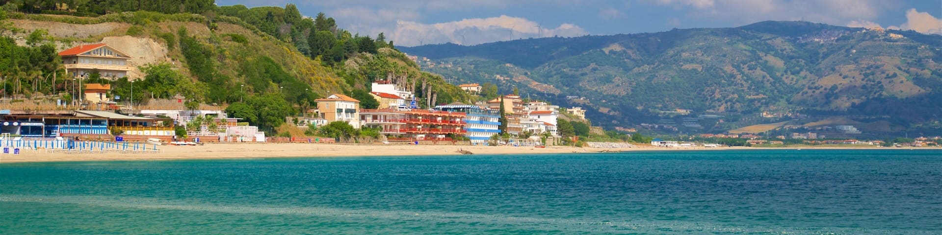 A coastal town with buildings along the beach, a clear blue sea, and mountains in the background.