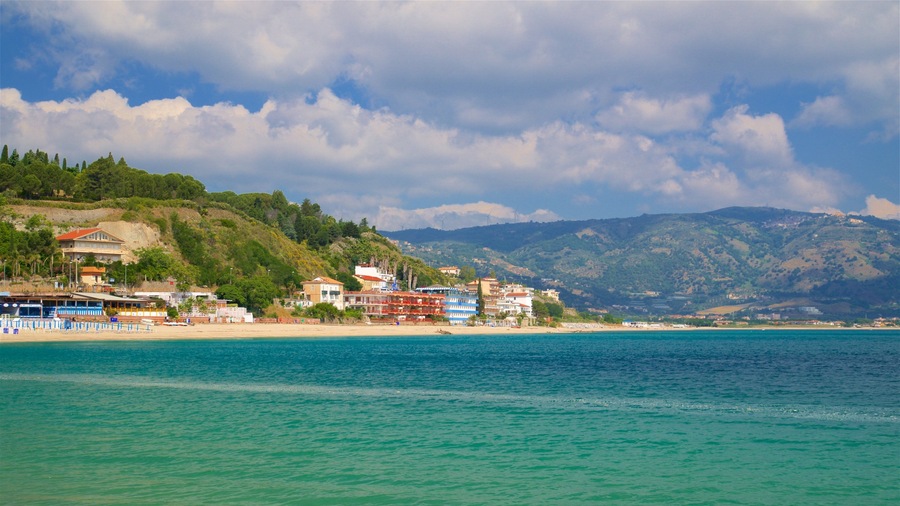 A coastal town with buildings along the beach, a clear blue sea, and mountains in the background.