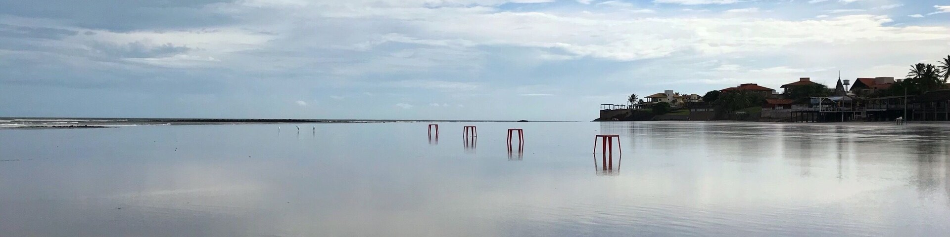 Uma praia tranquila com céu claro, algumas nuvens e uma linha de costa distante com edifícios.