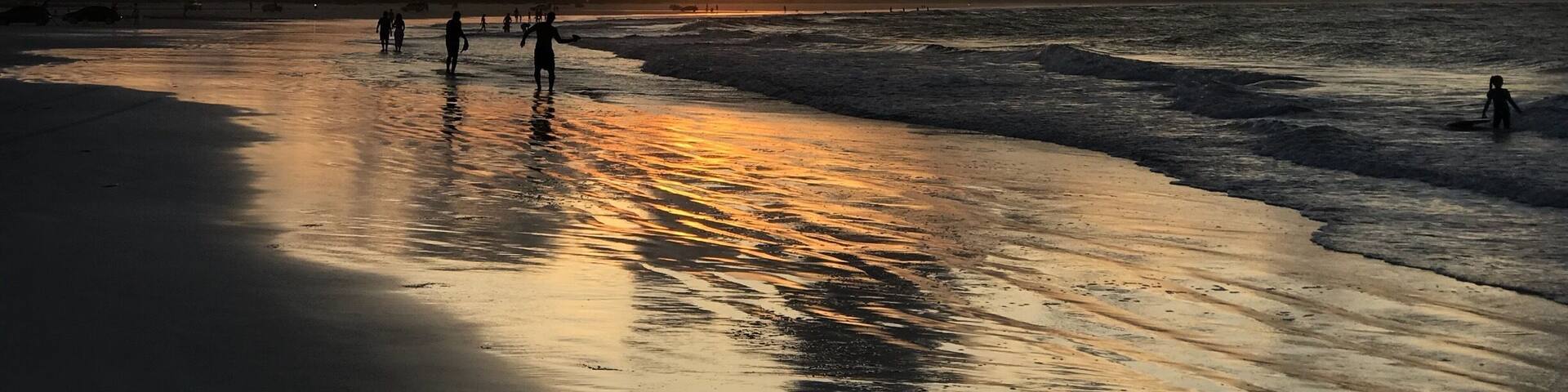 A beach at sunset with people walking along the shore and a sunset in the background.