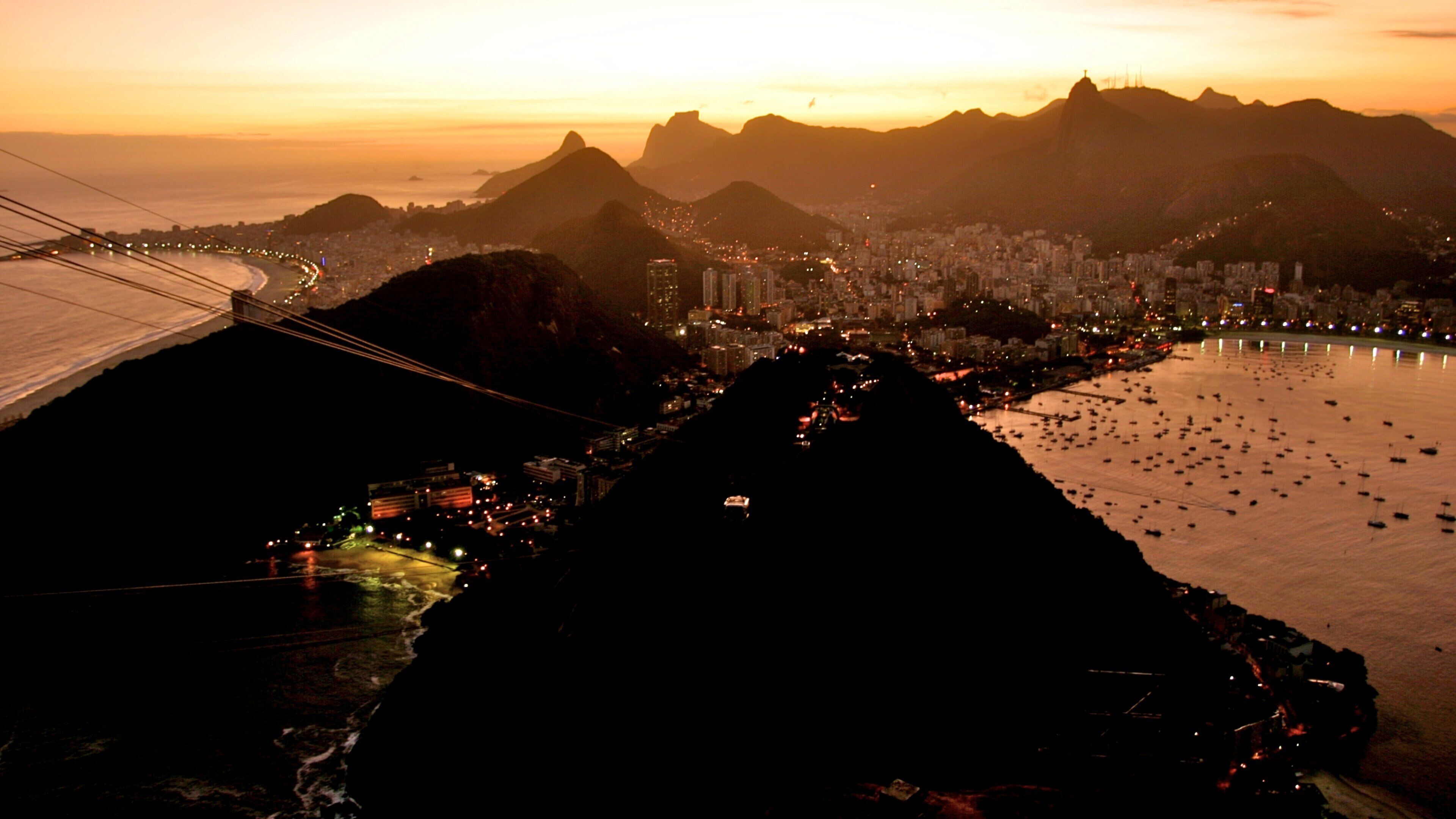 A cityscape at sunset with a prominent mountain range in the background and a river with numerous boats.