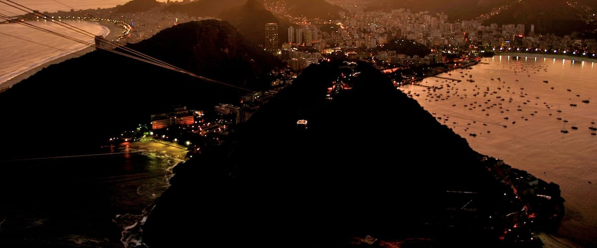A cityscape at sunset with a prominent mountain range in the background and a river with numerous boats.