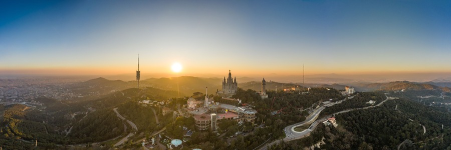 A panoramic view of a city with a prominent cathedral, a Ferris wheel, and winding roads amidst a hilly landscape.