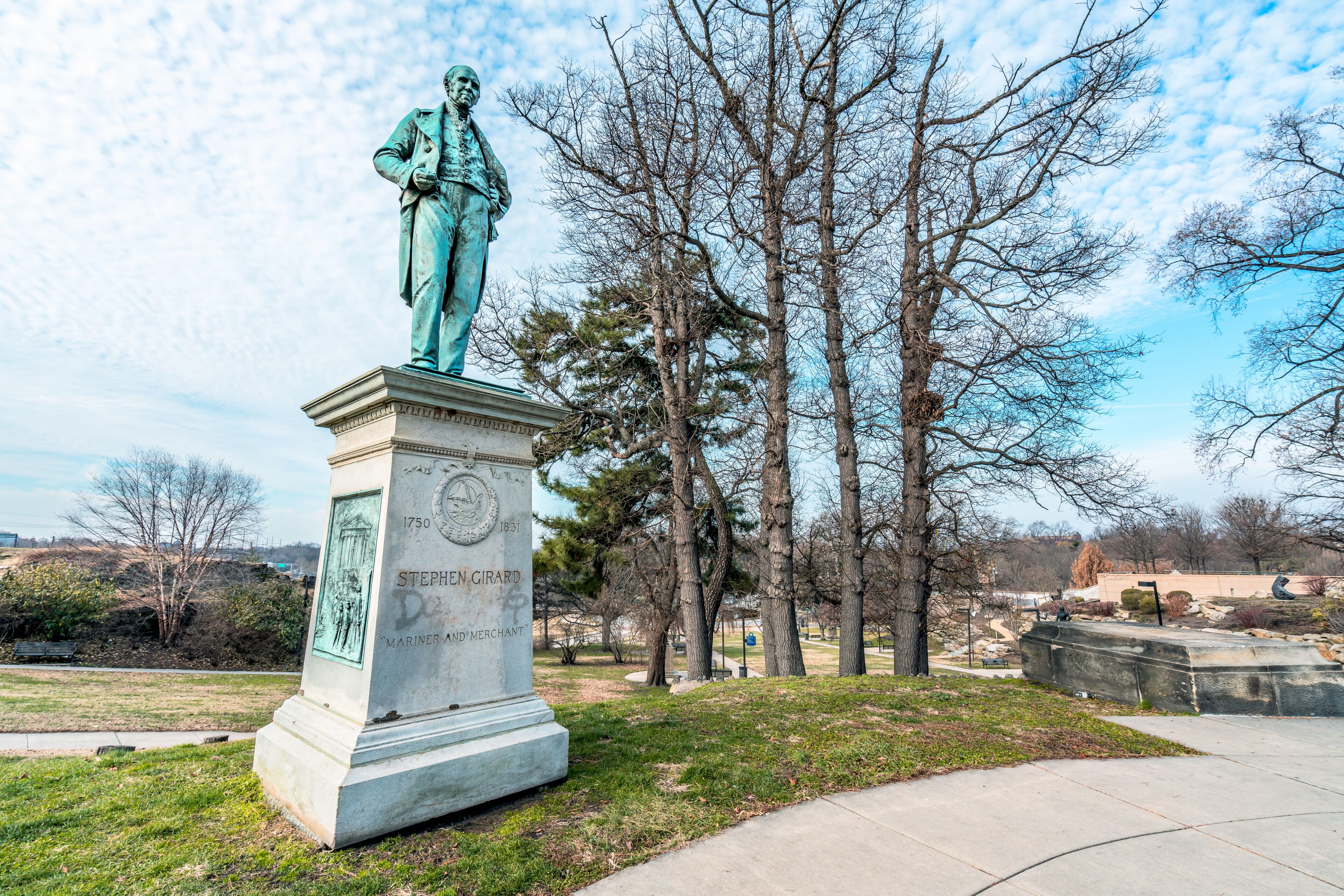A statue of Stephen Girard in a park with trees and a clear sky.