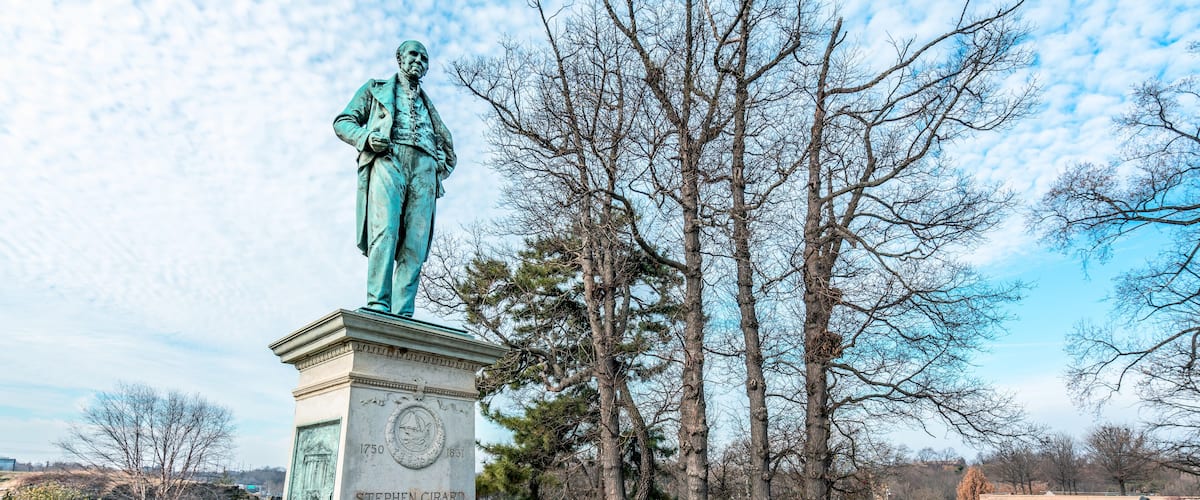 A statue of Stephen Girard in a park with trees and a clear sky.