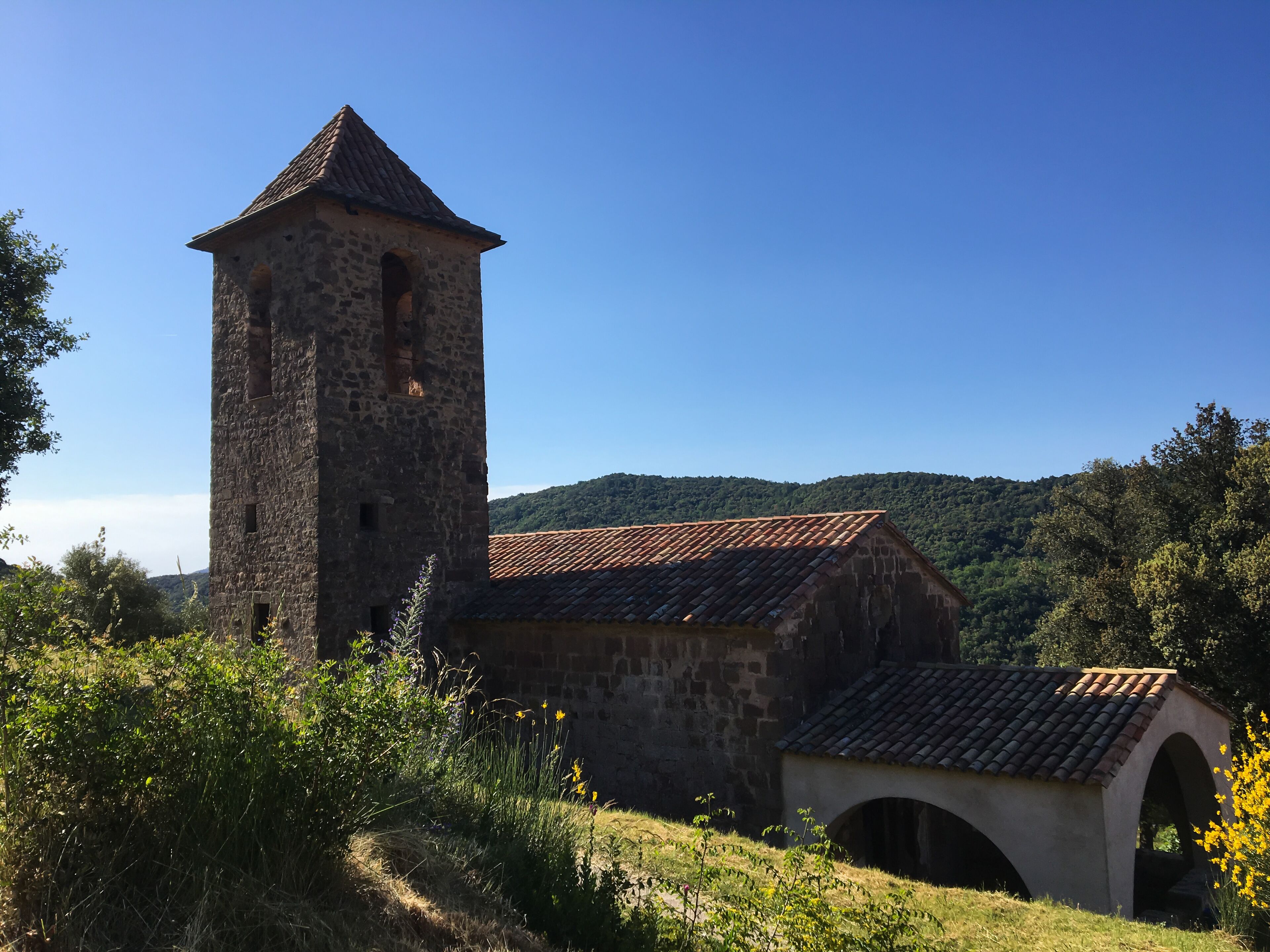 A stone tower with a tiled roof, a small building with a tiled roof, and an arched bridge in a hilly landscape.
