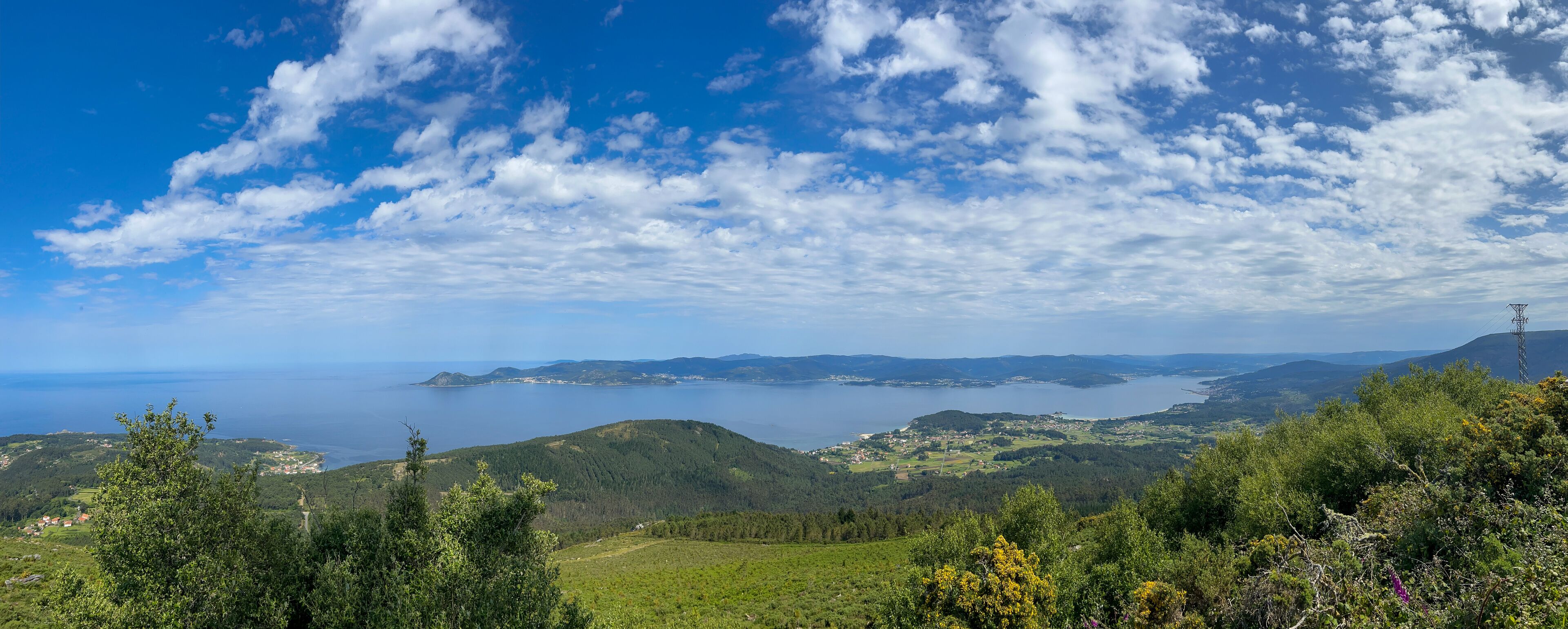 A stunning photo of Ria de Muros e Noia, Galicia, showcasing vast water, lush green hills, and quaint coastal towns. Fluffy clouds add drama to the serene landscape.