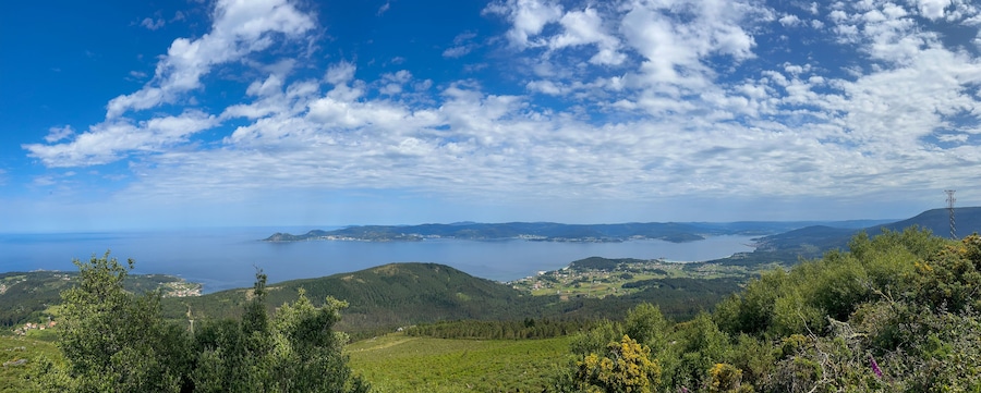 A stunning photo of Ria de Muros e Noia, Galicia, showcasing vast water, lush green hills, and quaint coastal towns. Fluffy clouds add drama to the serene landscape.