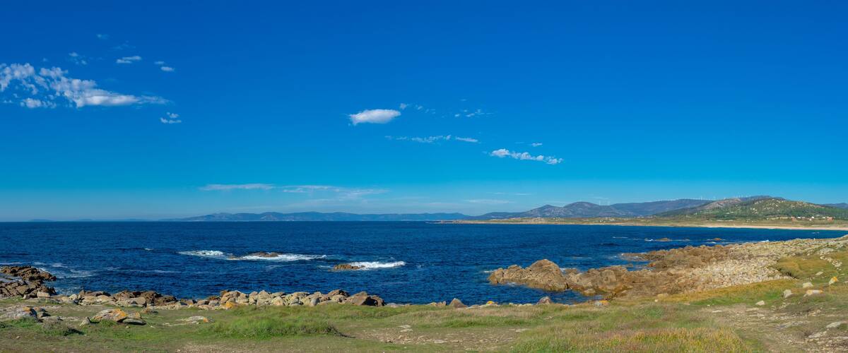 A coastal landscape with a rocky shoreline, a clear blue sky, and distant mountains.