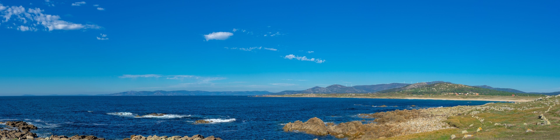 A coastal landscape with a rocky shoreline, a clear blue sky, and distant mountains.