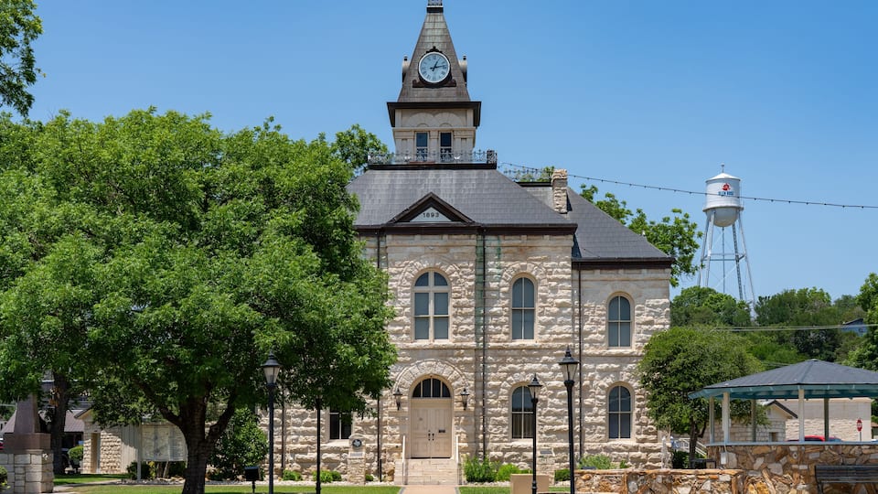 A historic stone church with a clock tower, surrounded by lush green trees and a clear blue sky.