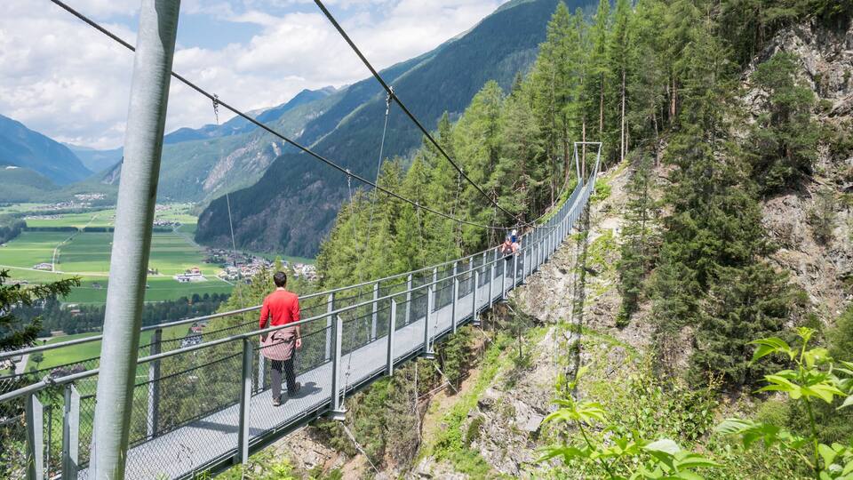 A suspension bridge with a person in a red shirt walking on it, surrounded by green mountains and a clear sky.