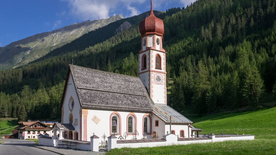 A church with a red dome and a clock tower, surrounded by a mountainous landscape and a green field.