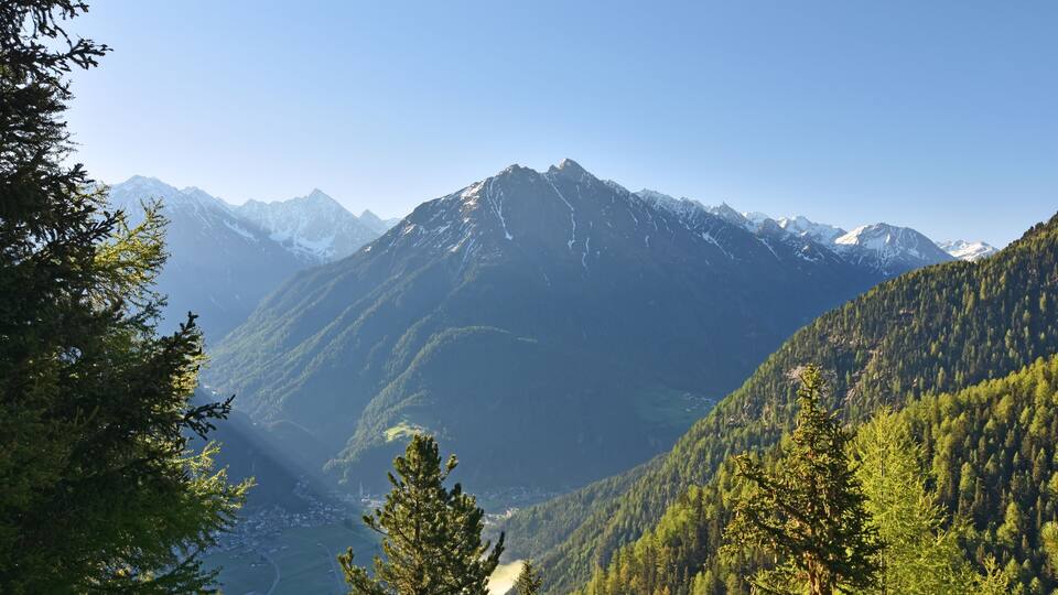 A mountain range with snow-capped peaks, dense forested valleys, and a clear sky.