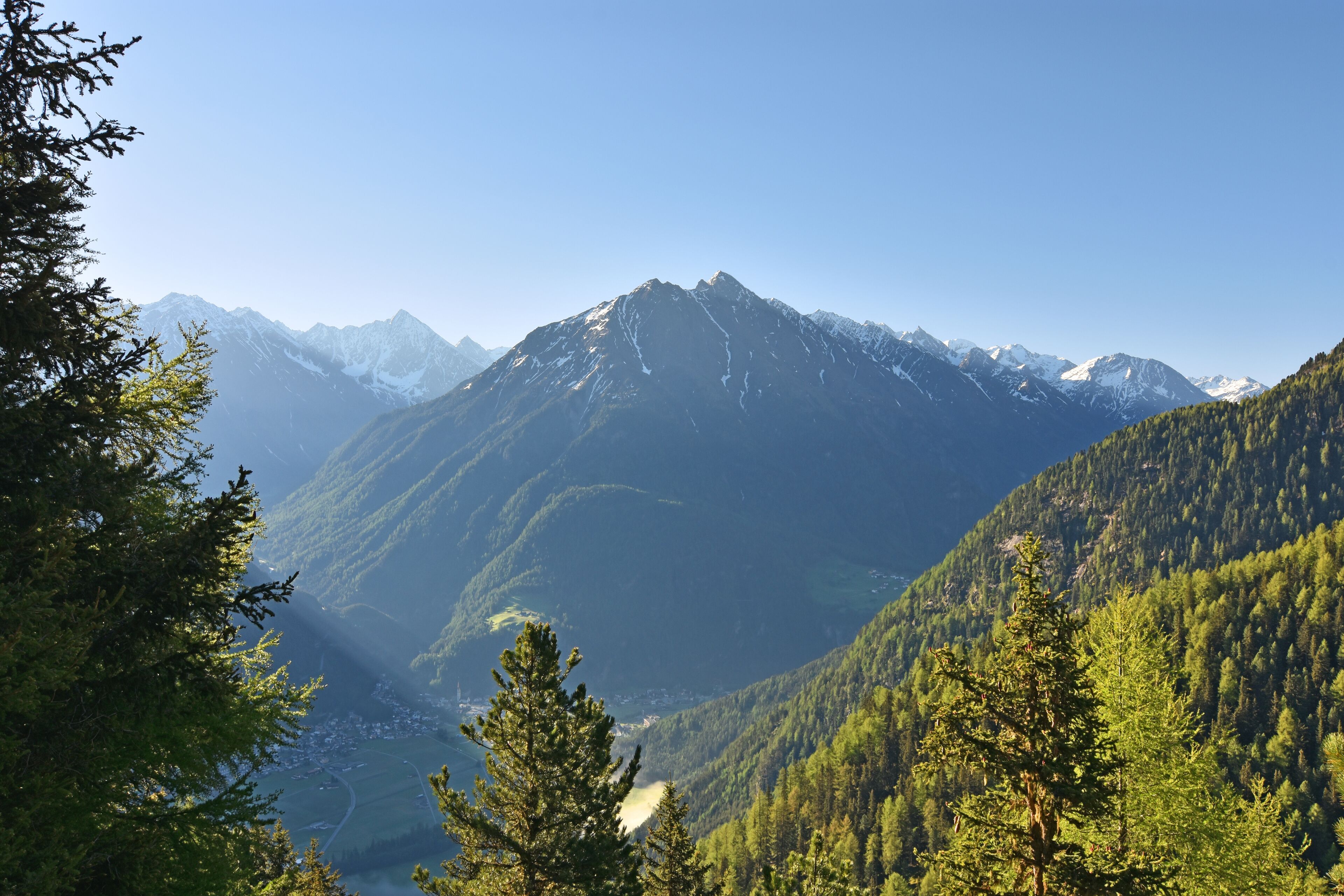 A mountain range with snow-capped peaks, dense forested valleys, and a clear sky.