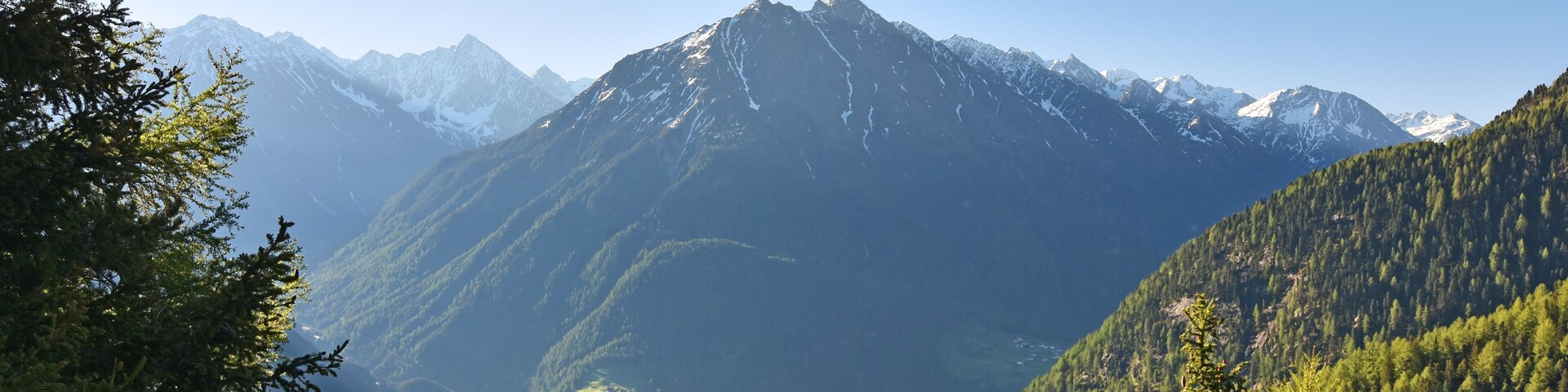 A mountain range with snow-capped peaks, dense forested valleys, and a clear sky.