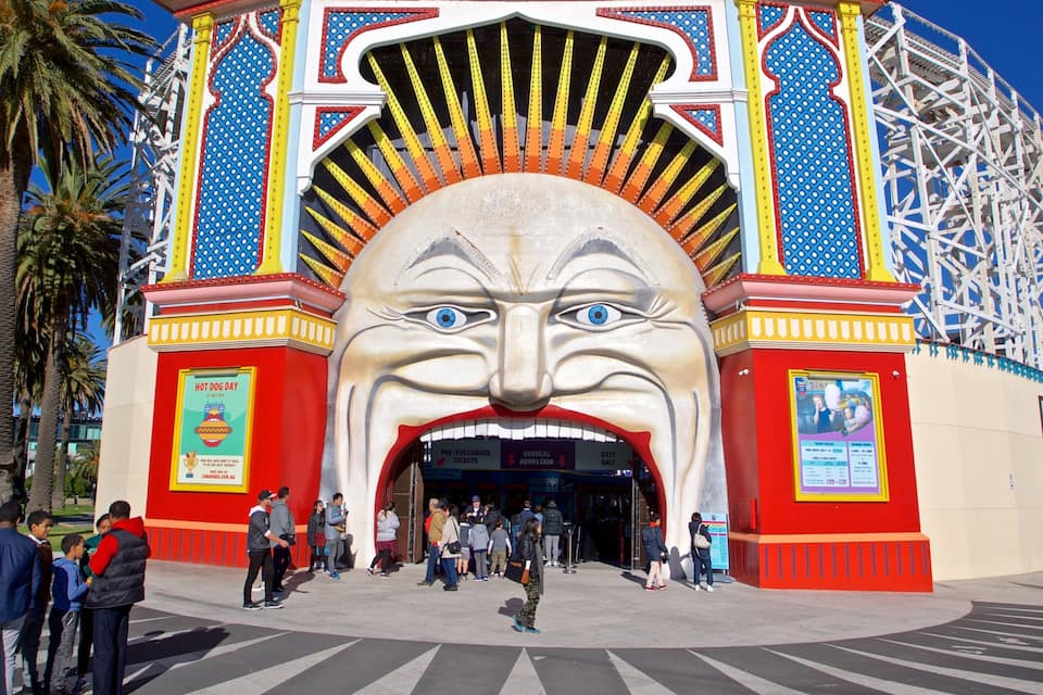 A colorful entrance with a large face sculpture, palm trees, and a roller coaster in the background.
