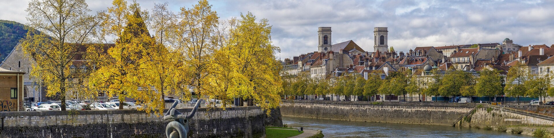 Un río con una escultura, árboles otoñales y edificios históricos al fondo.