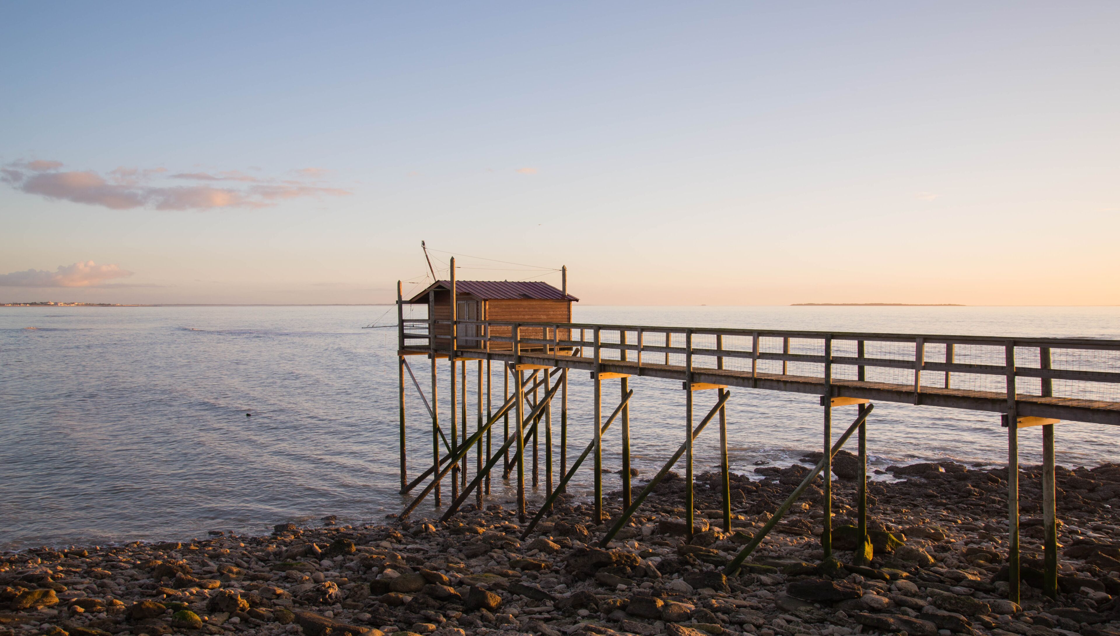 Un muelle de madera se extiende sobre aguas tranquilas, con una pequeña construcción de madera al final, y una costa rocosa.