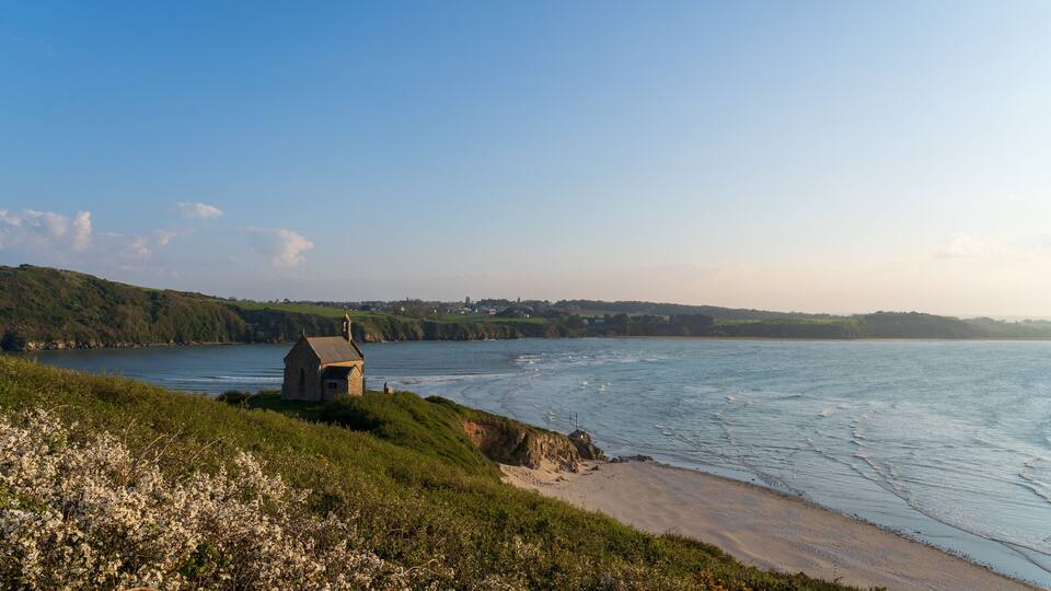 A coastal landscape with a small stone building near the shore, surrounded by greenery and a clear blue sky.