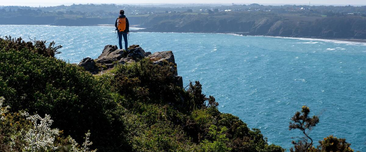 A person standing on a rocky cliff overlooking a coastal area with a clear view of the sea and distant hills.