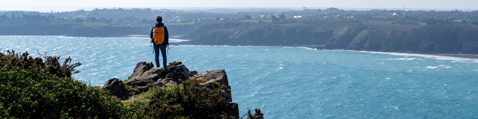 A person standing on a rocky cliff overlooking a coastal area with a clear view of the sea and distant hills.