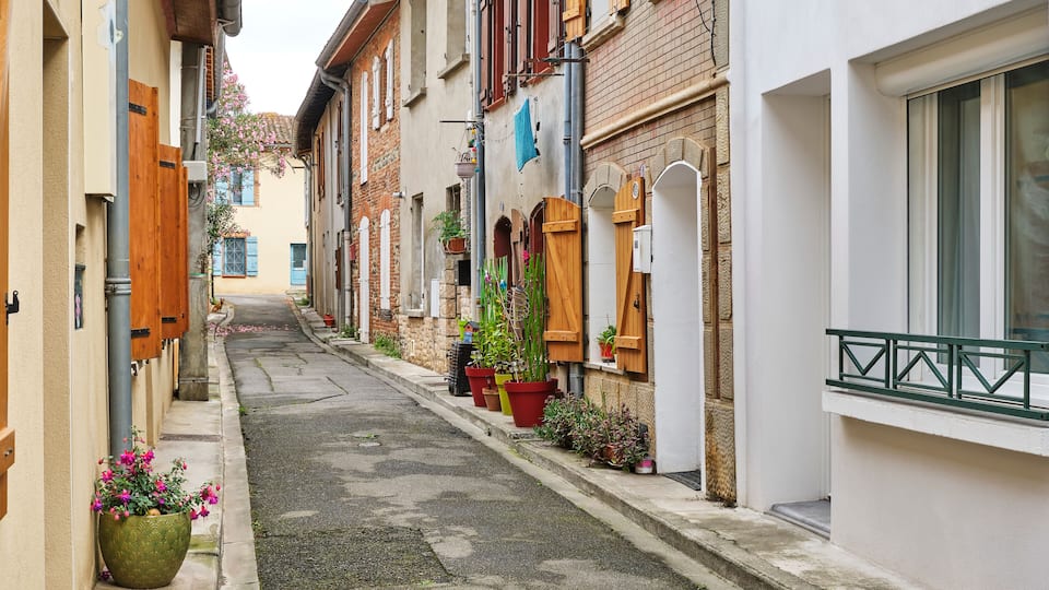 A narrow street lined with buildings, potted plants, and wooden shutters.