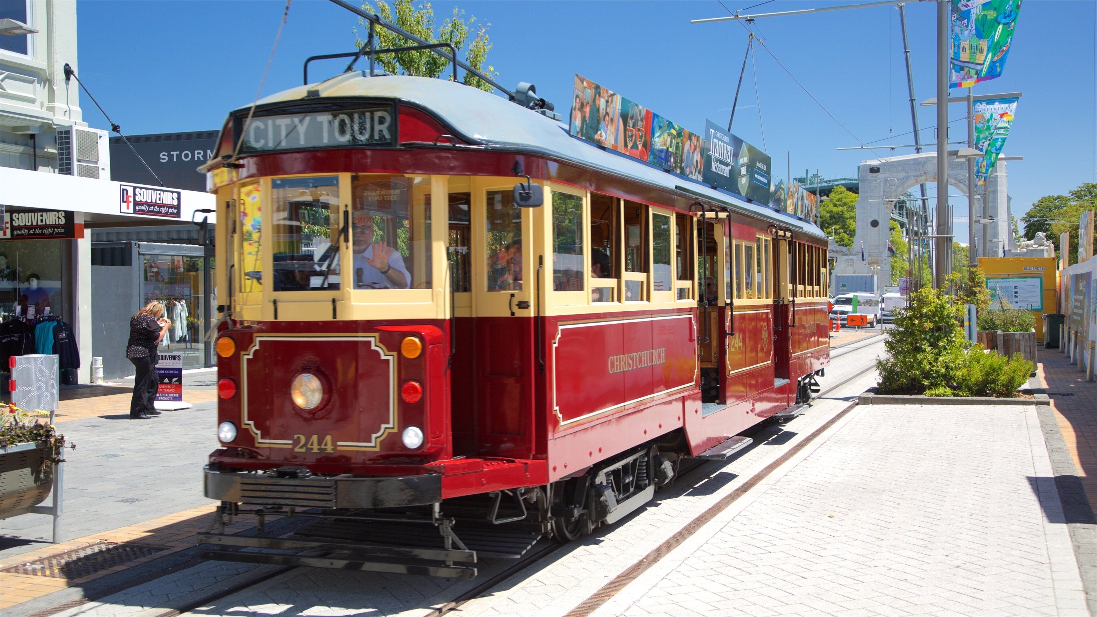 Eine rote und gelbe Straßenbahn mit dem Aufdruck "City Tour" fährt auf einer Gleisstrecke in einem städtischen Gebiet mit Geschäften und blauem Himmel.