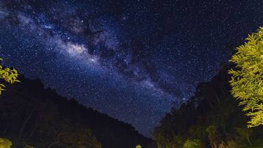 A starry night sky over a mountainous area with houses and trees.
