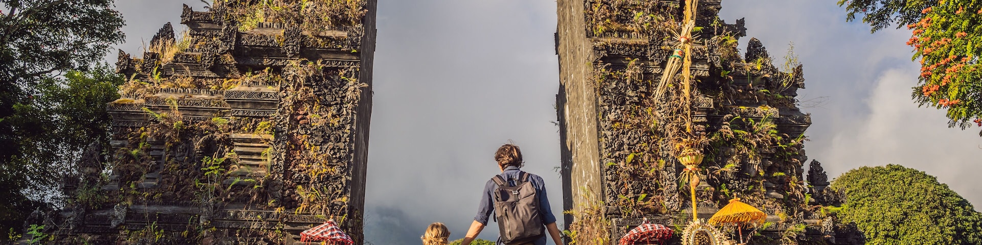 Um homem e uma criança caminham por portões de pedra antiga com esculturas detalhadas e um exuberante cenário verde.