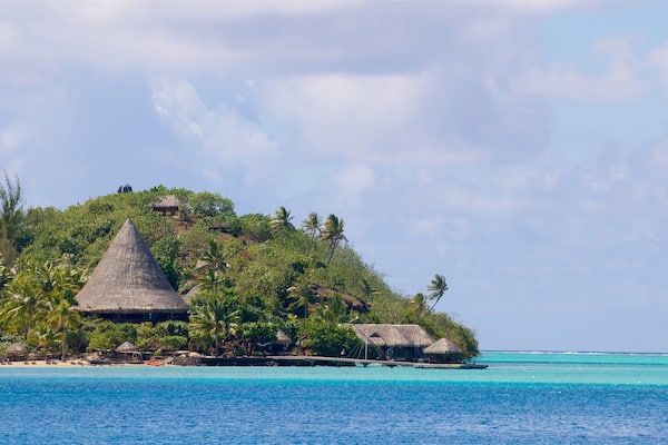 Une île tropicale avec des huttes aux toits de chaume, entourée de palmiers et bordée d’eaux d’un bleu cristallin.