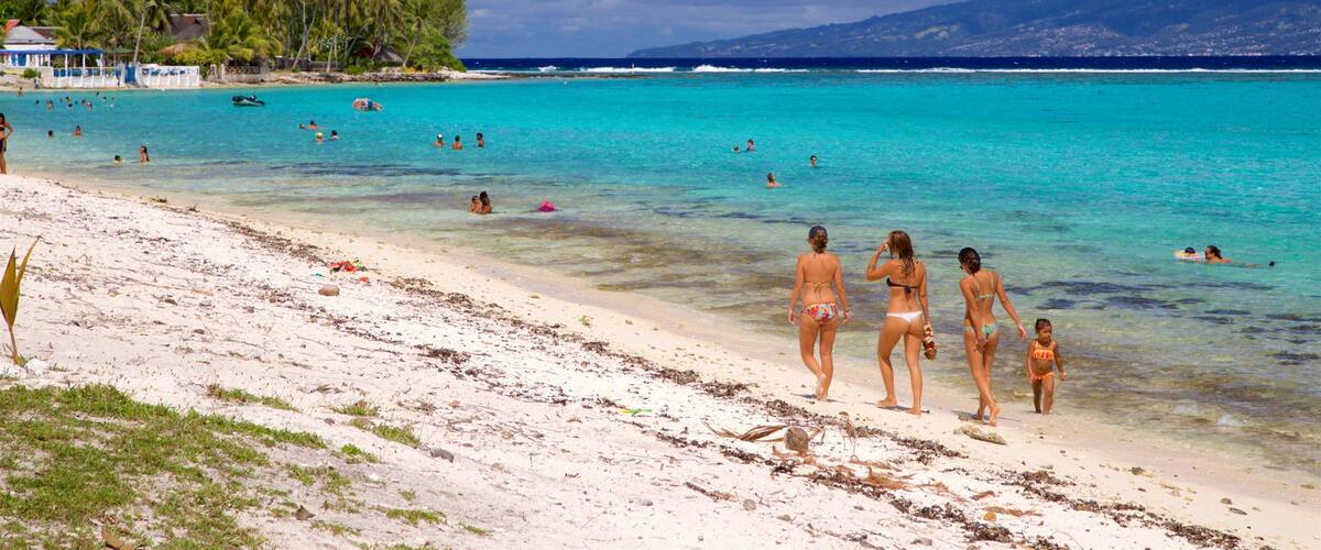 A beach with clear turquoise water, people swimming and walking on the shore, and a backdrop of palm trees and distant mountains.