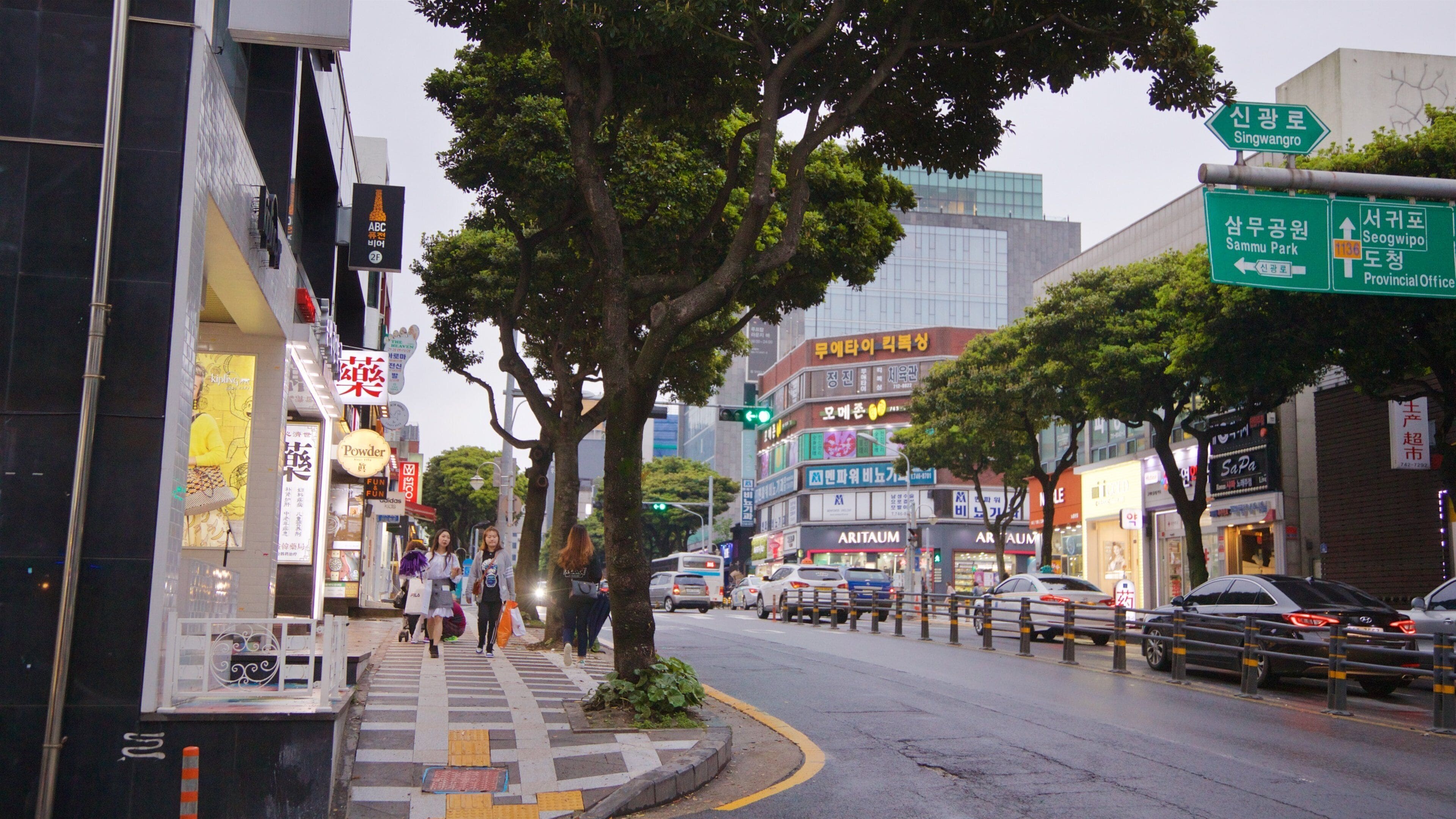Una calle urbana concurrida, con peatones, automóviles y diversos rótulos comerciales.