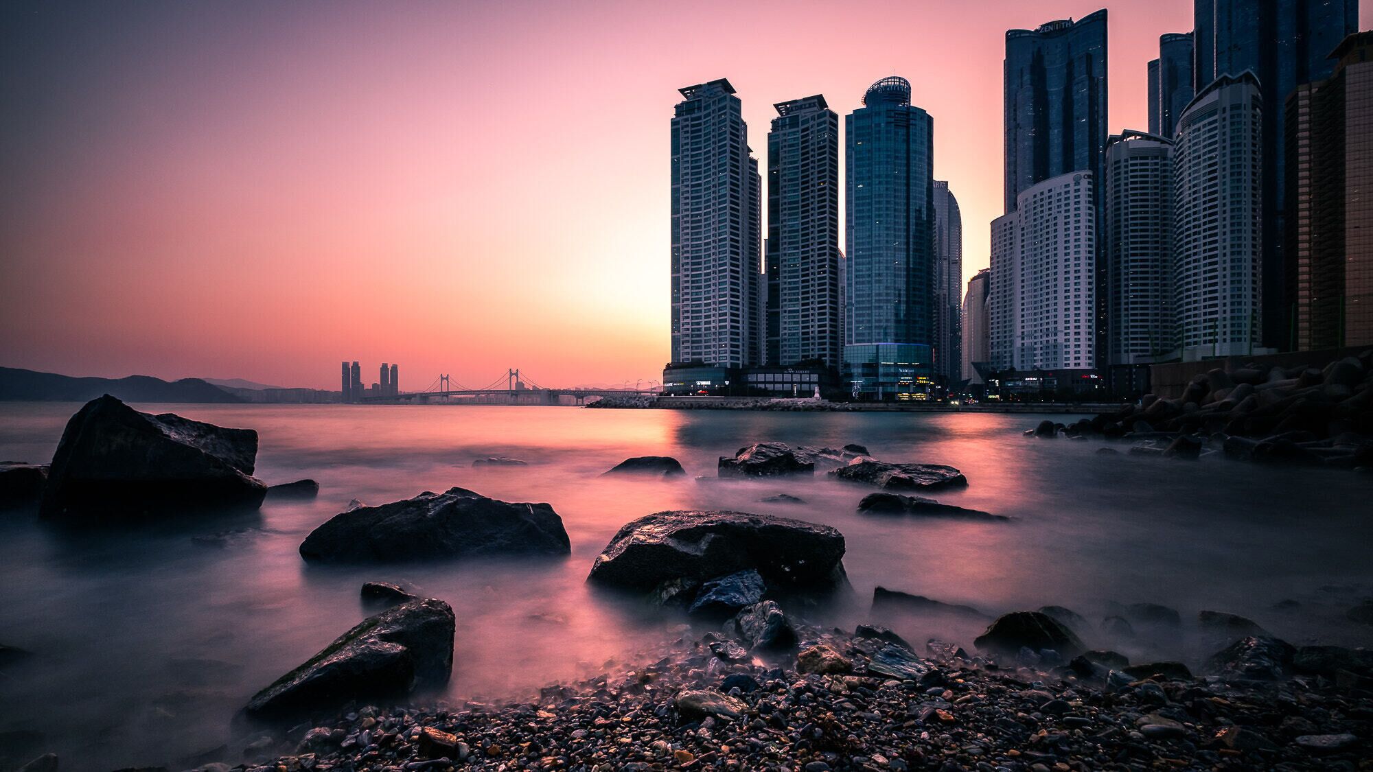 Un paisaje urbano al atardecer con un río en primer plano y rascacielos al fondo.