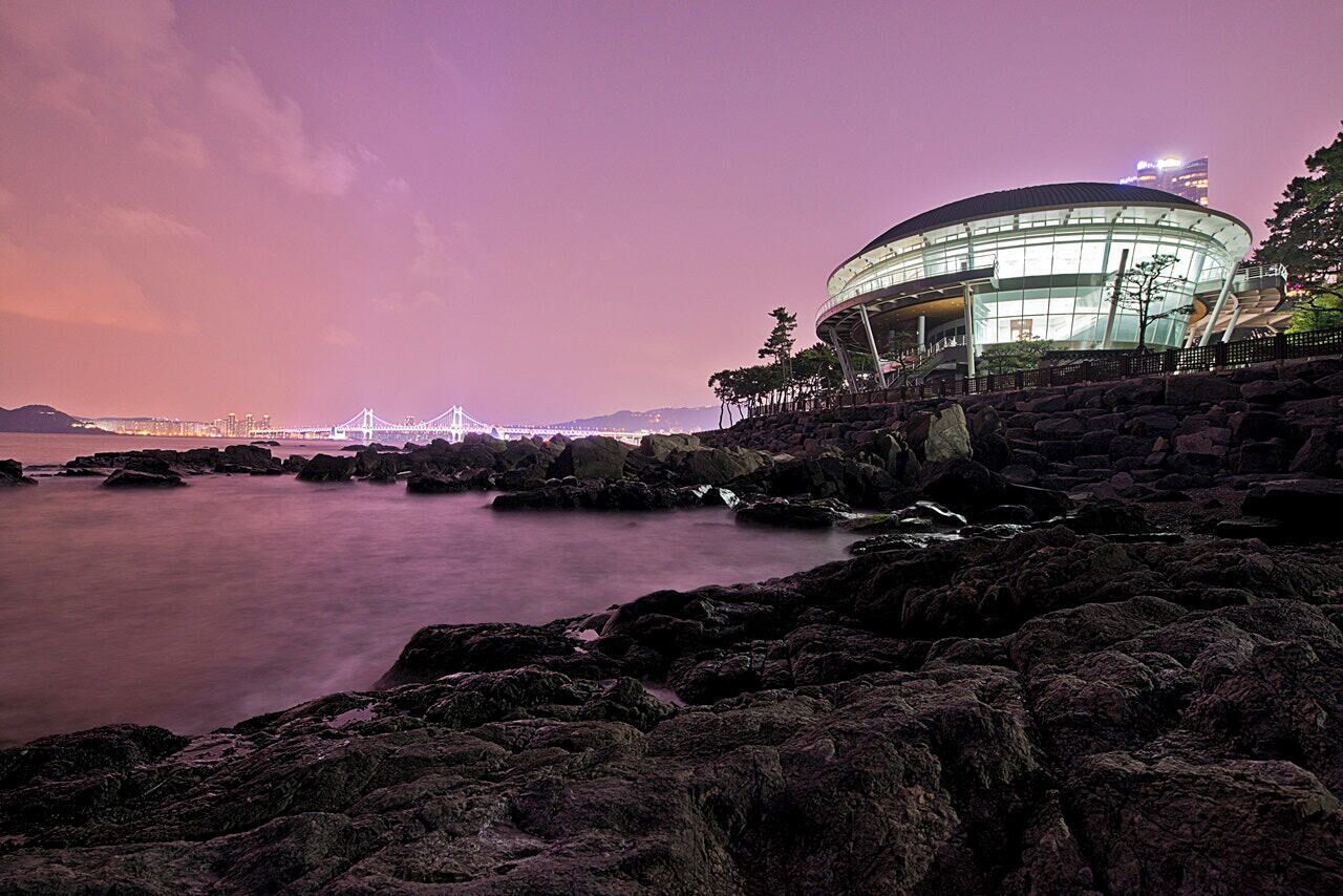 A coastal scene with a modern building, rocky shoreline, and a bridge in the background.