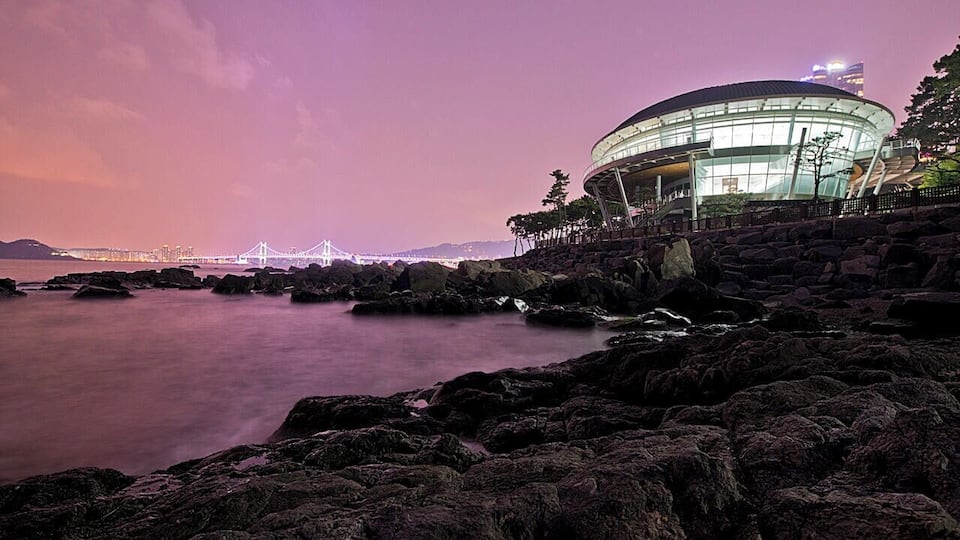 A coastal scene with a modern building, rocky shoreline, and a bridge in the background.
