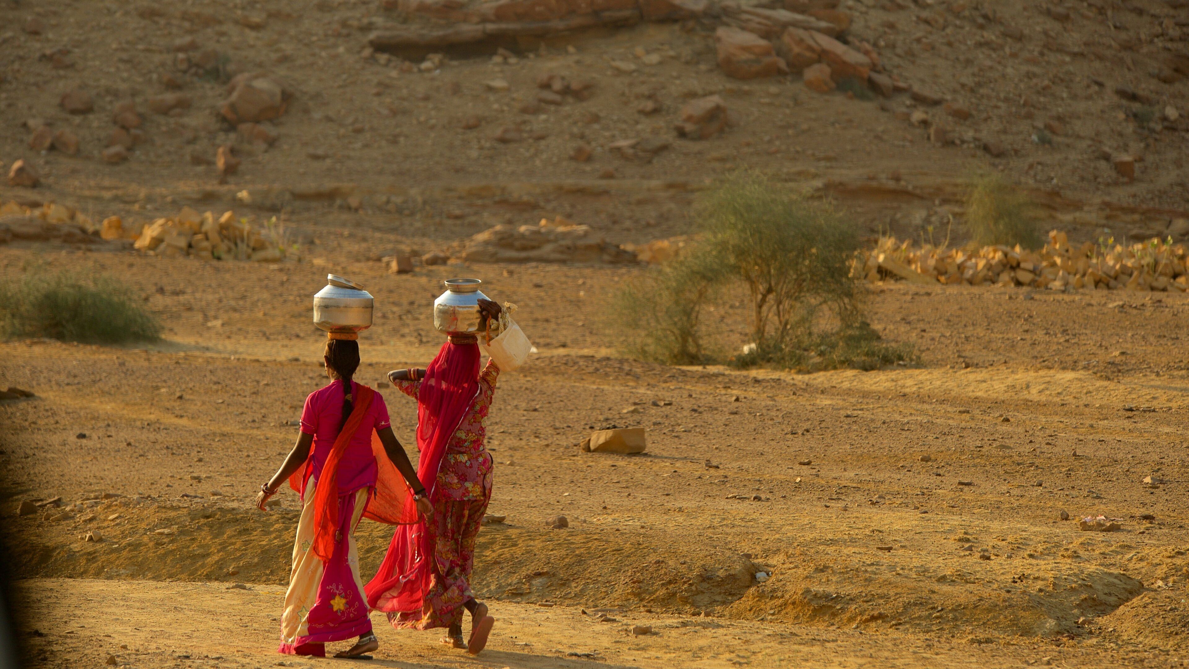 Two women walking in a desert with rocky terrain and sparse vegetation.
