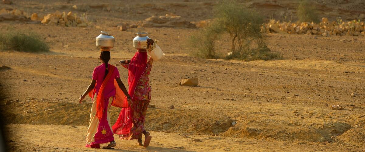 Two women walking in a desert with rocky terrain and sparse vegetation.