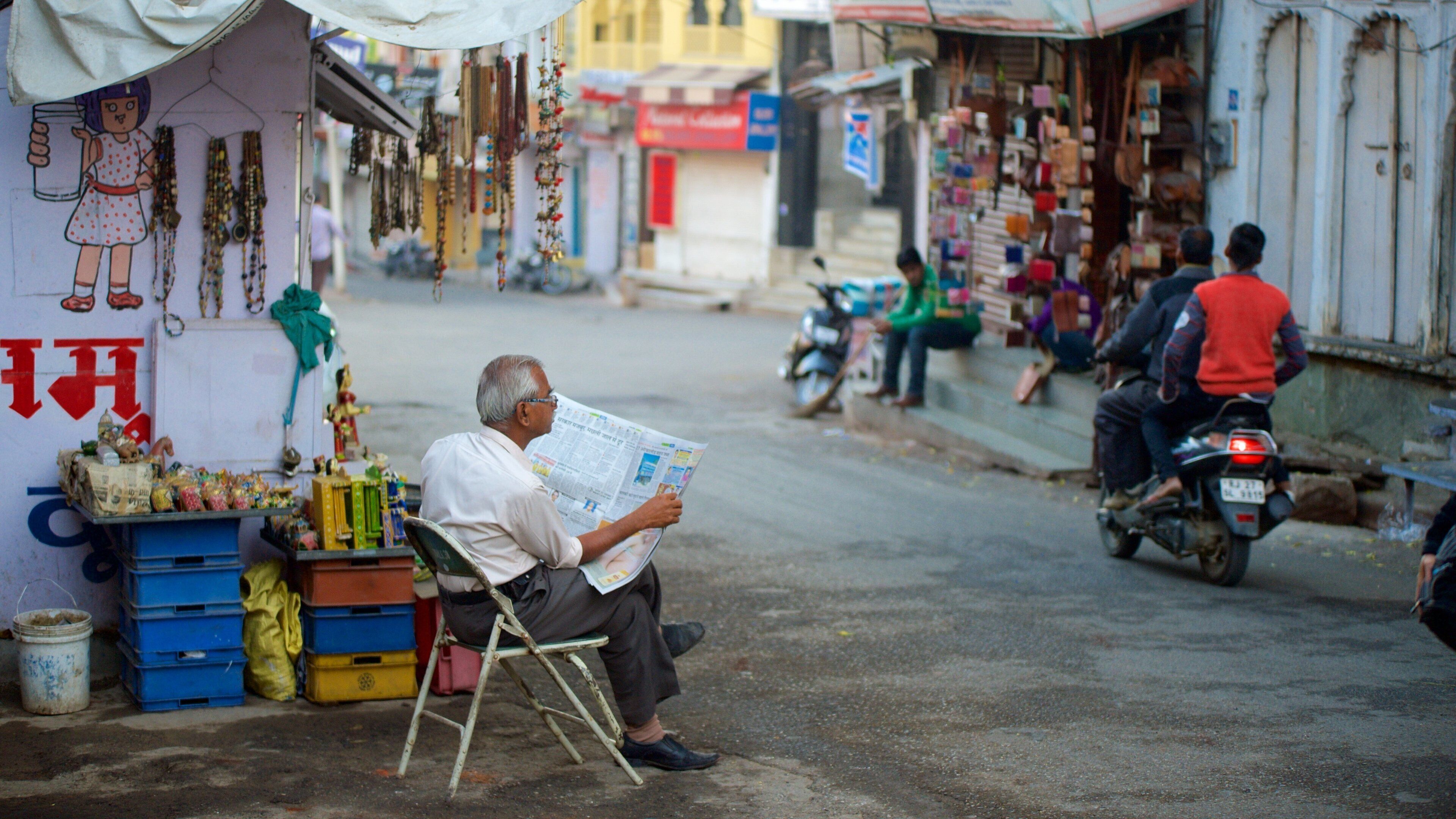 Cena de rua com um homem lendo jornal, uma mulher em vestido pendurada numa parede e diversas lojas.